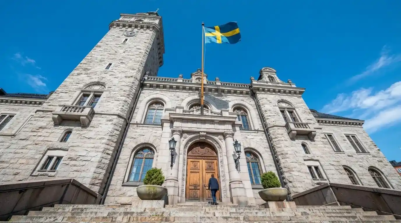 Ett gammalt stadshus i sten fotograferat underifrån mot en klarblå himmel, med den svenska flaggan som fladdrar ovanför entrén.