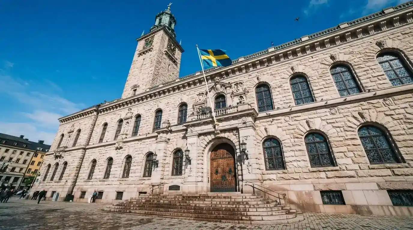 Ett gammalt stadshus i sten fotograferat underifrån mot en klarblå himmel, med den svenska flaggan som fladdrar ovanför entrén.